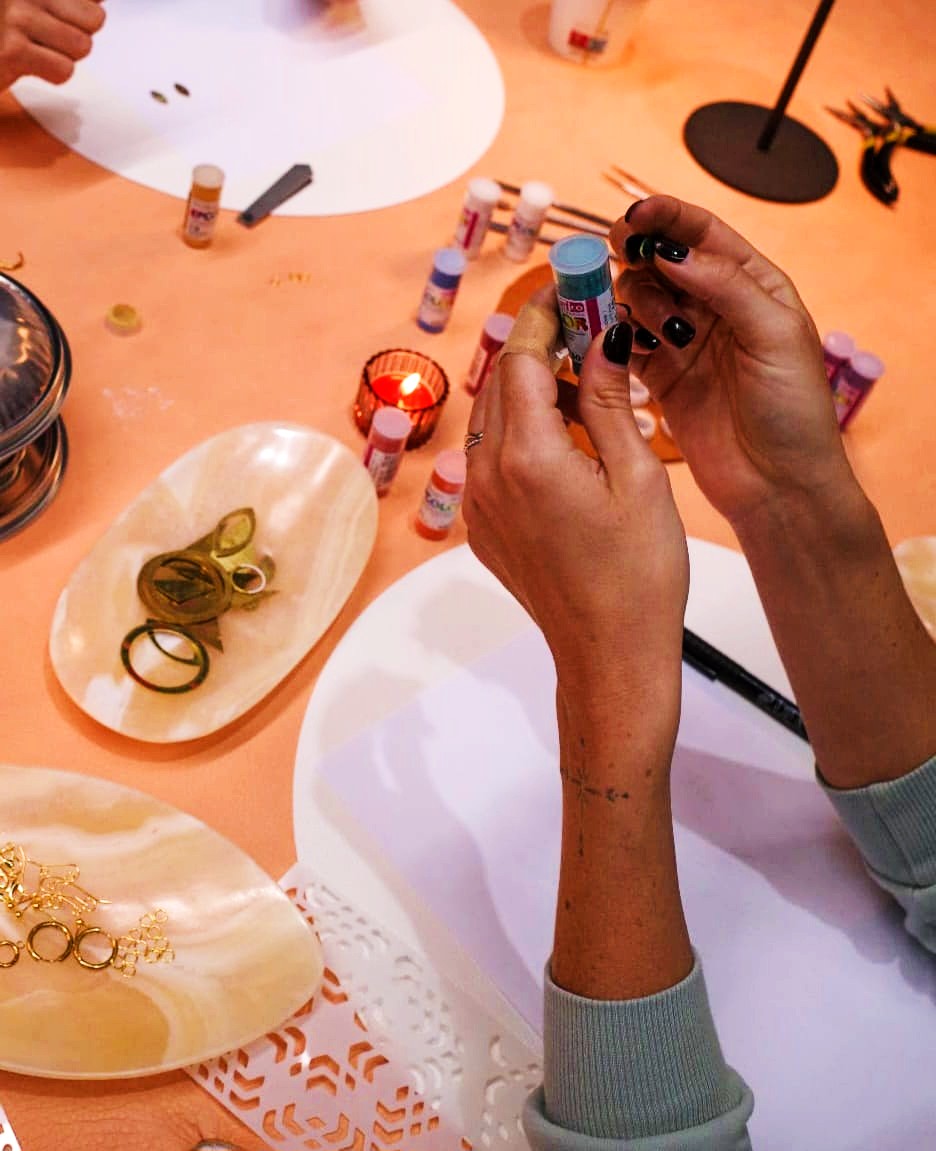 A close-up of a person's hands holding a small tube with a blue cap, surrounded by enamel colours, rings, jewelleryand decorative trays on a peach-colored table.